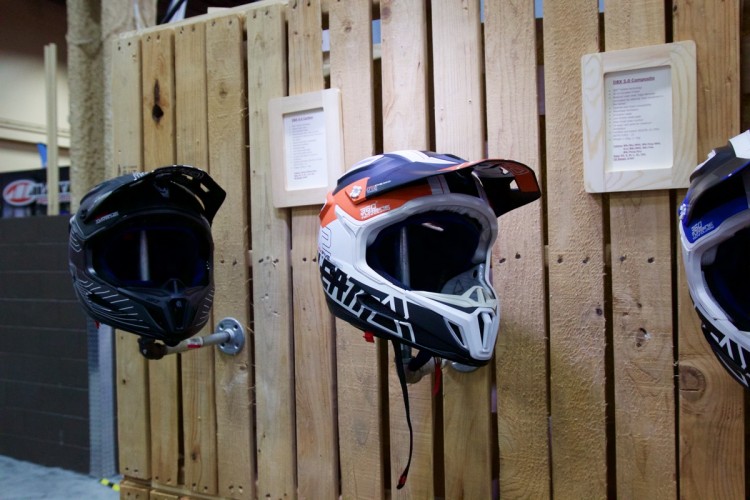 Two motorcycle helmets displayed on a wooden wall, one black and one white with orange and blue accents. A wooden sign in the background provides information about helmet specifications.