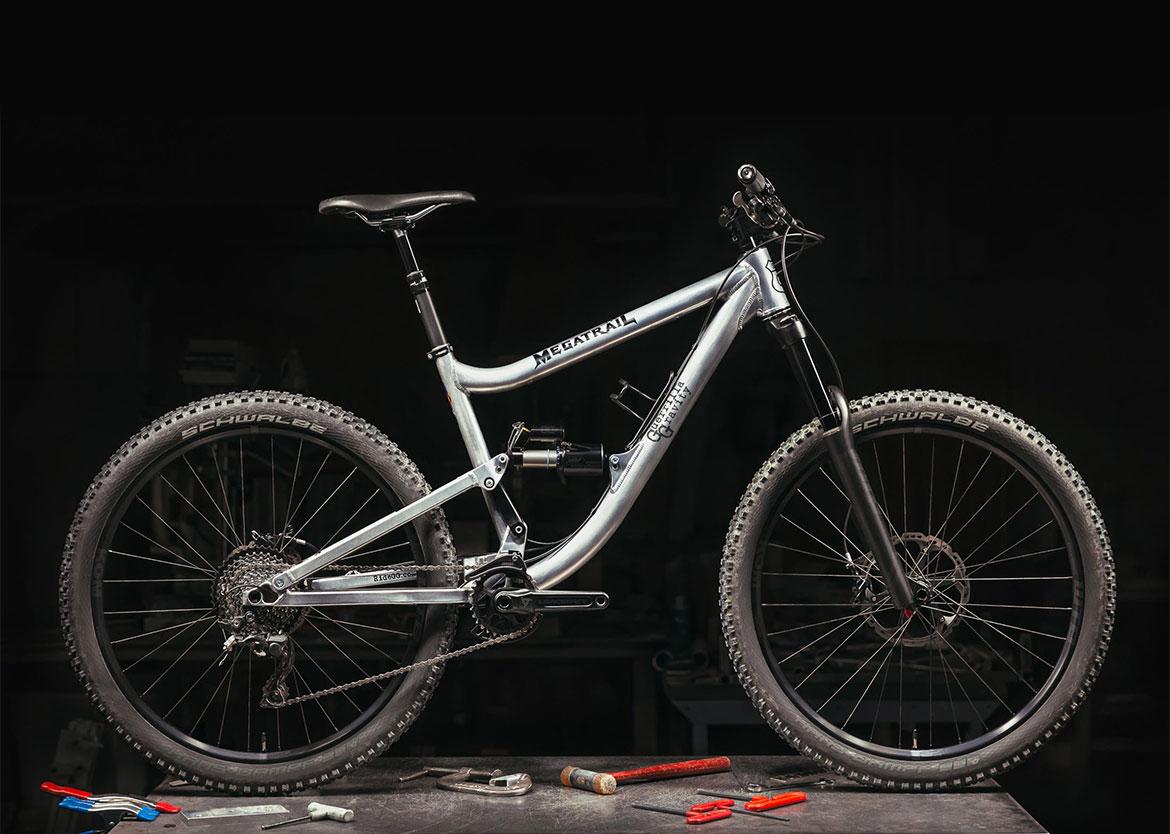Guerrilla Gravity Megatrail: A polished silver mountain bike with wide tires and a modern frame design, positioned on a dark workbench surrounded by various tools. The bike features a suspension system, disc brakes, and a prominent brand label on the frame.