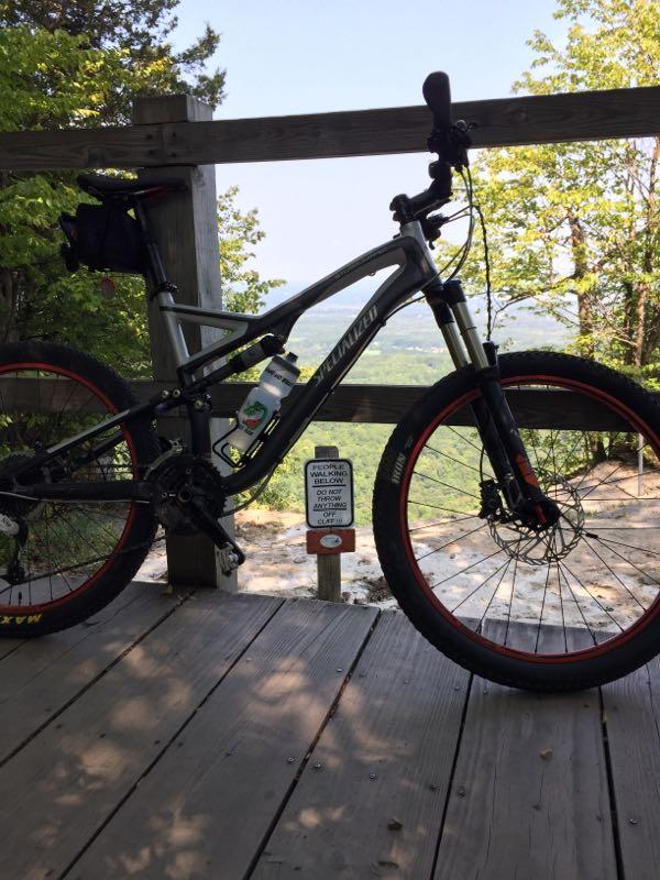 Alt text: A mountain bike is parked on a wooden deck, overlooking a scenic view with trees in the foreground. A warning sign is visible nearby, indicating safety precautions. The bike has red accents and a water bottle attached. Thatcher State Park mountain bike trail.