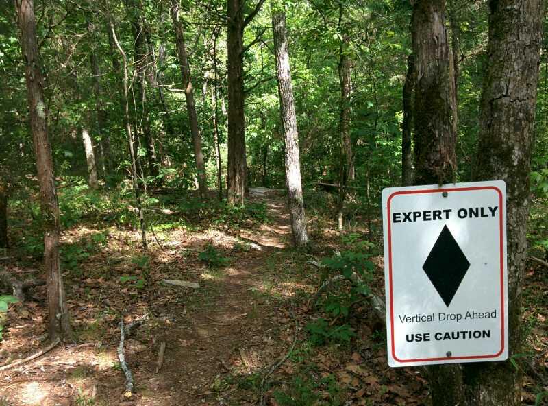A wooded trail with a warning sign nearby that reads "EXPERT ONLY" with a diamond symbol and the message "Vertical Drop Ahead, Use Caution." The scene features tall trees and a winding path, indicating a potentially challenging hiking area. Clinton Nature Preserve mountain bike trail.