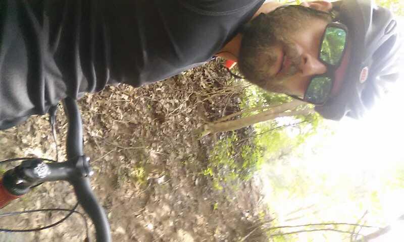 A person wearing a cycling helmet and sunglasses is taking a selfie while riding a bicycle on a dirt trail surrounded by trees. The setting appears to be a natural environment with leaves and greenery in the background. Saluda Shoals Park mountain bike trail.