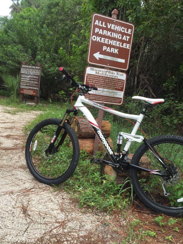 A mountain bike parked on a trail near a sign that reads "All Vehicle Parking at Okeeheelee Park" with an arrow pointing left. The surrounding area is lush with greenery and a dirt pathway. Additional signs about mountain bike trail modifications are visible in the background. Okeeheelee Park / Pinehurst / Green Acres Freedom Park mountain bike trail.
