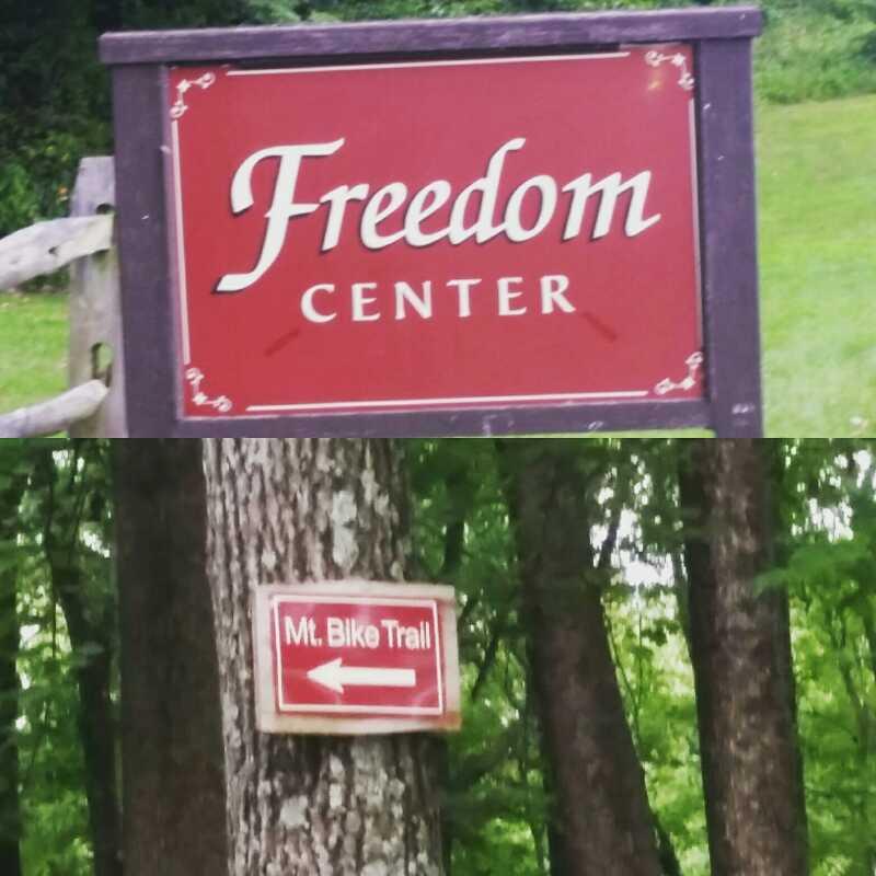 A large sign reading "Freedom Center" at the top, with a secondary sign pointing left that says "Mt. Bike Trail." The background features a forested area with lush greenery. Freedom Center mountain bike trail.
