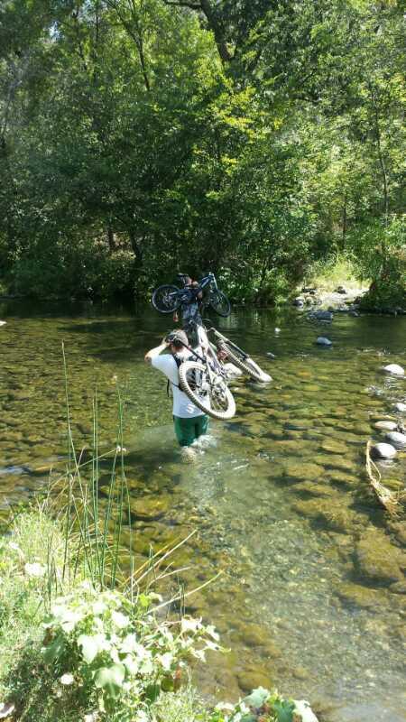 A person wading through a shallow, clear stream while carrying two bicycles on their shoulders. Lush greenery surrounds the area, with rocks visible in the water and sunlight filtering through the trees. Upper Bidwell Park mountain bike trail.