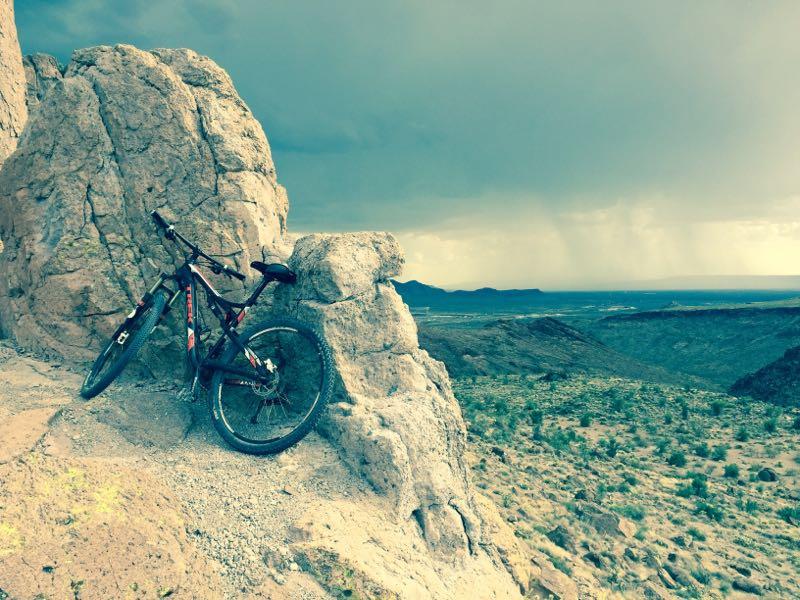A mountain bike leaning against a large rock formation, with a vast landscape visible in the background under a moody sky. The scene suggests an adventurous outdoor setting, possibly after or before a rain shower. Cerbat Foothills Recreation Area mountain bike trail.