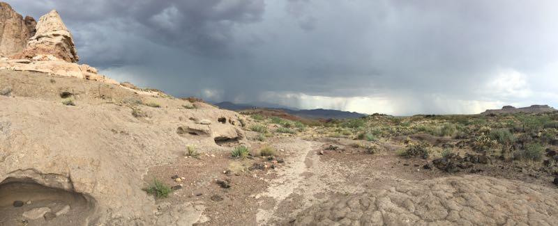 A panoramic view of a rugged landscape featuring rocky formations under a dramatic sky. Dark clouds loom over distant mountains, hinting at an approaching storm. The foreground includes dry, cracked earth interspersed with patches of green vegetation and scattered stones. Cerbat Foothills Recreation Area mountain bike trail.