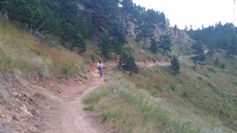 A person riding a bicycle along a winding dirt trail surrounded by trees on a hillside. The scene captures the natural landscape with grassy areas and rocky outcrops, under a partly cloudy sky. HLMP mountain bike trail.