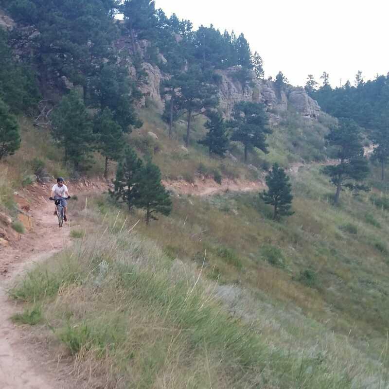 A person riding a bicycle on a winding dirt trail surrounded by trees and rocky hills, with a natural landscape in the background. HLMP mountain bike trail.