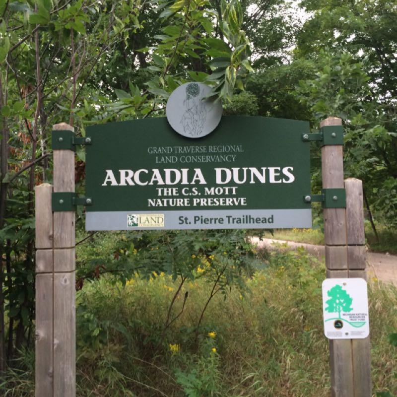 Sign marking the entrance to the Arcadia Dunes, C.S. Mott Nature Preserve, managed by the Grand Traverse Regional Land Conservancy. The sign indicates the St. Pierre Trailhead and features lush greenery in the background. Arcadia Dunes mountain bike trail.