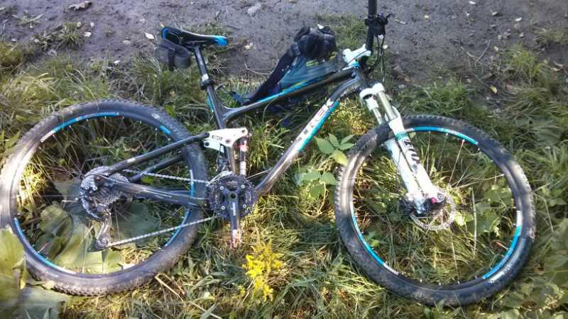 A mountain bike lying on the ground, surrounded by grass and wild plants. The bike features thick tires and a black frame with blue accents. A small backpack is positioned on the bike. The background is a mixture of dirt and greenery. Hydrocut mountain bike trail.