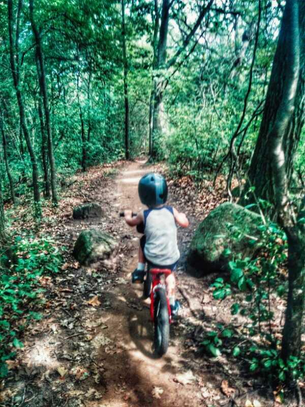 A young child riding a small red bicycle down a dirt path surrounded by trees and greenery. The child is wearing a helmet and is focused on navigating the trail, which features rocks and leaves scattered along the way. Kettle Moraine John Muir + Emma Carlin mountain bike trail.