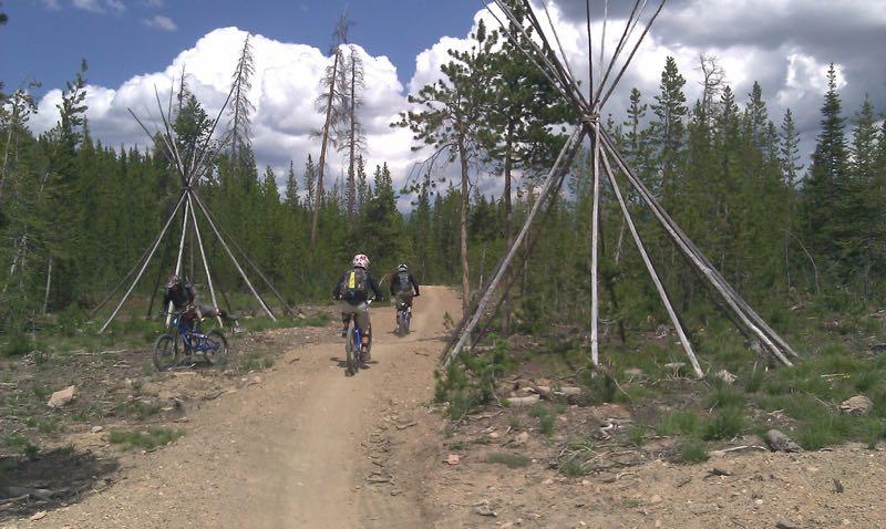 Three mountain bikers ride along a dirt trail surrounded by tall trees and wooden structures resembling tipis. The sky is partly cloudy, adding a sense of openness to the serene outdoor setting. Trestle Bike Park mountain bike trail.