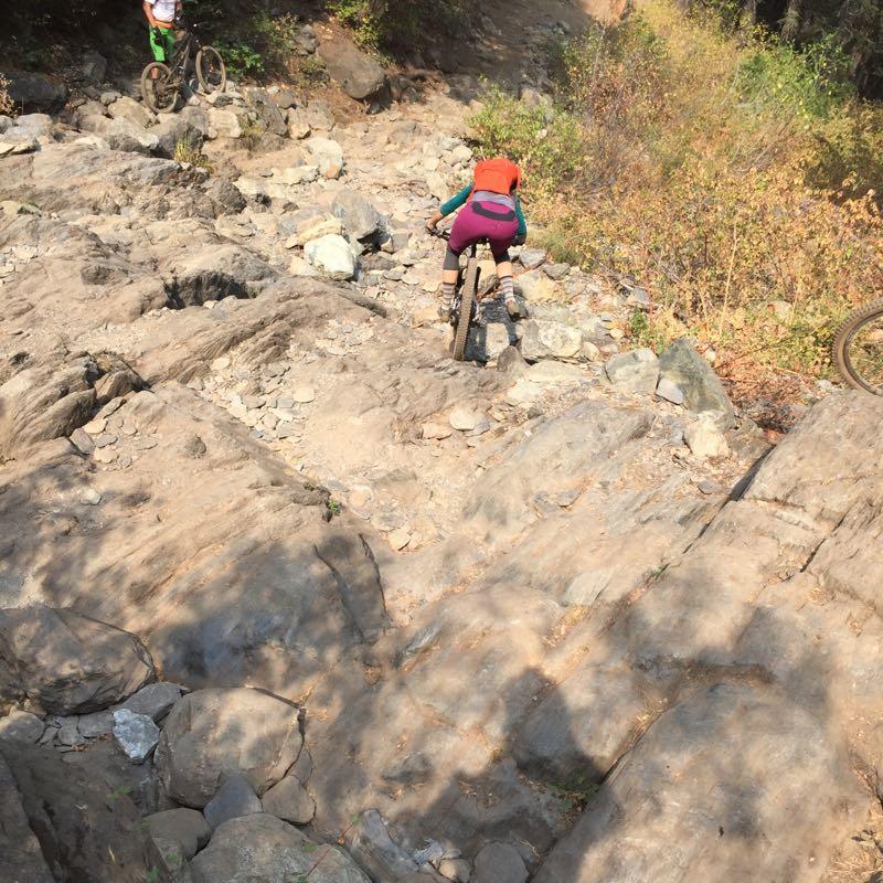 Two mountain bikers navigate a rocky terrain. One rider, in a bright orange top and purple shorts, is riding down a steep, uneven surface covered in stones. The second rider is visible in the background, closer to the top of the slope, wearing a white shirt and green shorts. The scene is set in a natural outdoor environment with greenery surrounding the rocky path. Downieville Downhill mountain bike trail.