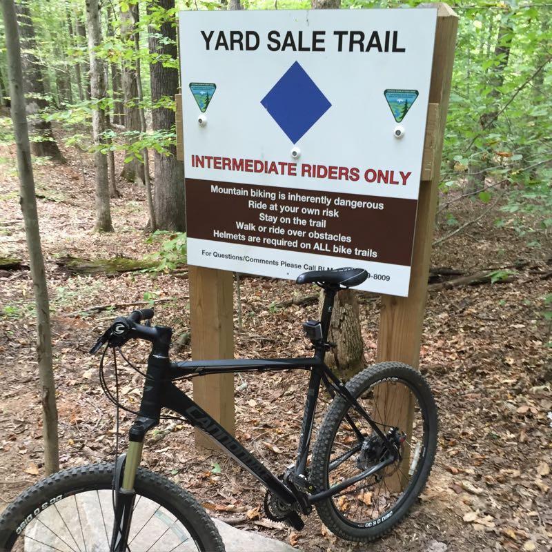 A black mountain bike is leaning against a wooden sign that reads "YARD SALE TRAIL" with a blue diamond symbol. The sign indicates it is for "INTERMEDIATE RIDERS ONLY" and includes safety warnings about mountain biking. The background shows a forested area with green foliage and fallen leaves on the ground. Meadowood mountain bike trail.