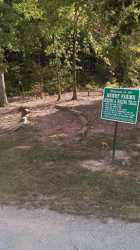 A gravel path leads into a wooded area, with a signpost on the right that reads "Welcome to the Henry Farms Hiking & Biking Trail." The sign provides information about the trail and its features. The surrounding area is lush with greenery and trees, indicating a natural setting. Henry Farm Park mountain bike trail.