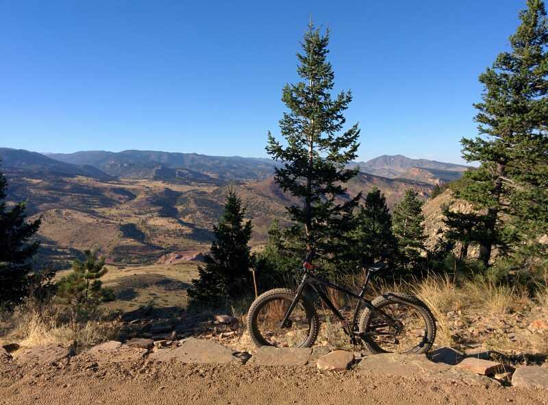 A mountain bike is resting against a tree on a rocky outcrop, overlooking a scenic valley with rolling hills and distant mountains under a clear blue sky. Pines and other vegetation are visible in the foreground, adding to the natural beauty of the landscape. Heil Valley Ranch mountain bike trail.