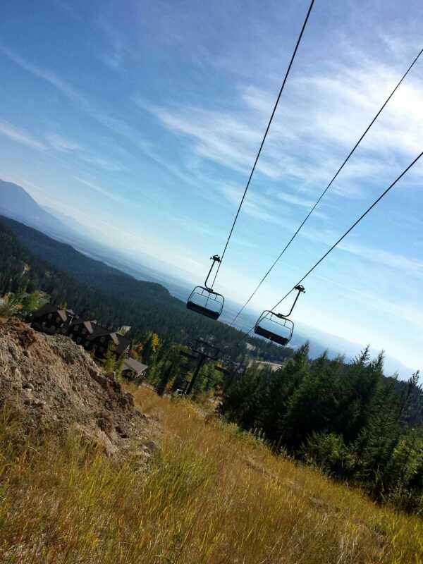 A scenic view of a mountain landscape featuring ski lift chairs suspended over grassy terrain, with a backdrop of trees and distant mountains under a clear blue sky. Whitefish Mountain Resort mountain bike trail.