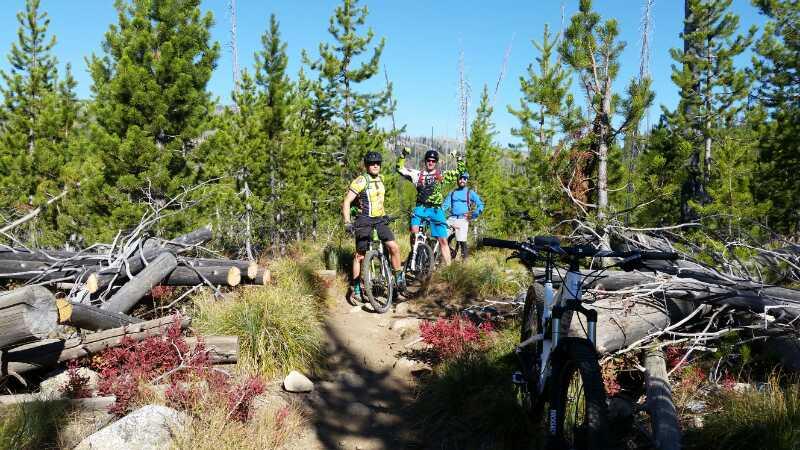Three mountain bikers pose on a wooded trail surrounded by tall pine trees and scattered logs. They are wearing helmets and cycling gear, with two bikes parked nearby. The sun is shining, creating a bright and vibrant atmosphere. Twenty Mile Creek Trail mountain bike trail.