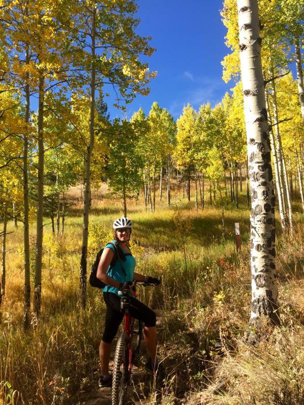 A cyclist in a turquoise shirt and helmet stands next to a red mountain bike on a dirt trail surrounded by vibrant yellow autumn trees and green grass under a clear blue sky. Golden Gate Canyon State Park mountain bike trail.