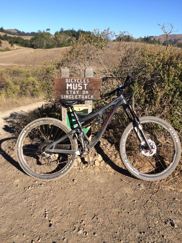 A mountain bike leaning against a wooden sign that reads "Bicycles Must Stay on Singletrack," set against a backdrop of a sunny, grassy landscape and trees. The bike features wide tires and a sturdy frame, indicating it's designed for off-road cycling. Camp Tamarancho mountain bike trail.