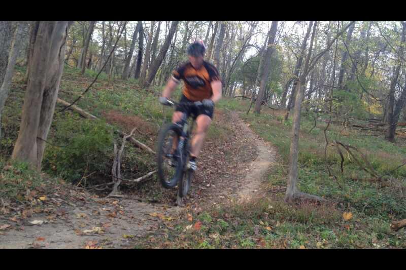 A person riding a mountain bike performs a jump over a small gap on a dirt trail surrounded by trees and autumn foliage. The rider is wearing an orange and black cycling jersey and a helmet. The image captures the dynamic motion of the jump in a natural outdoor setting. Capital View mountain bike trail.