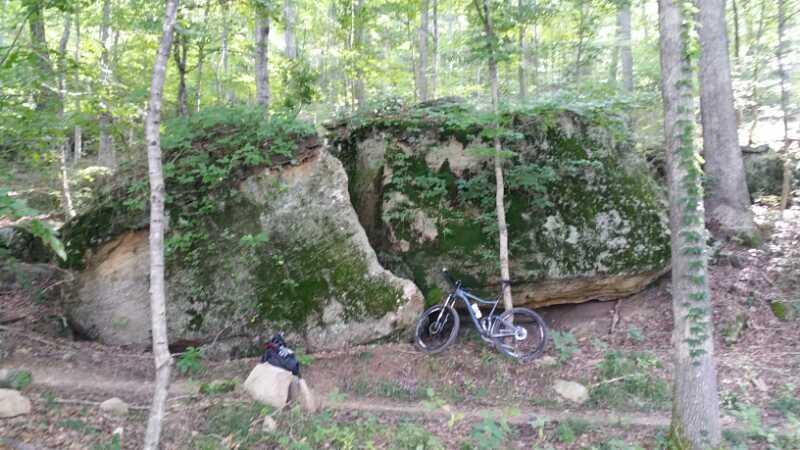 A large moss-covered rock with a partially cracked surface, surrounded by trees in a forested area. A mountain bike is propped against the rock, with a small backpack resting on the ground nearby, indicating a recreational outdoor setting. Brier Creek mountain bike trail.