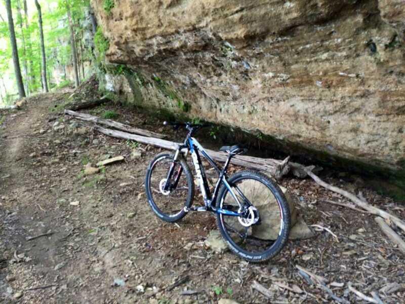 A mountain bike leaning against a large rock formation along a dirt trail surrounded by trees. Brier Creek mountain bike trail.