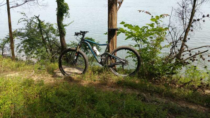 A mountain bike resting on a dirt path beside a body of water, surrounded by lush greenery and trees. The sun casts dappled shadows on the ground, creating a serene outdoor scene. Brier Creek mountain bike trail.