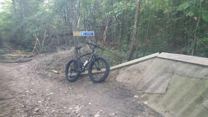 A black fat tire mountain bike is parked beside a dirt ramp on a trail surrounded by trees. A sign indicating the trail is visible in the background, with a chain preventing access. The ground is covered in leaves and dirt, typical of a wooded biking area. Horry County Bike Run Park mountain bike trail.