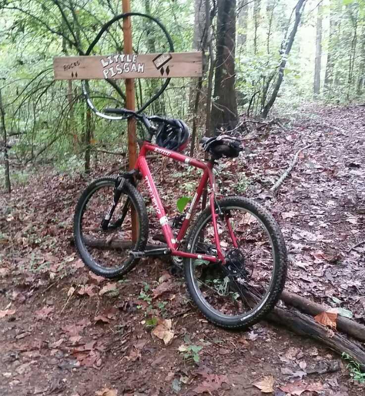 A red mountain bike leaning against a wooden sign that reads "Little Pisgah" in a wooded area, with a mix of dirt and fallen leaves on the ground. The background features dense trees and a trail leading into the woods. Hobby Park mountain bike trail.