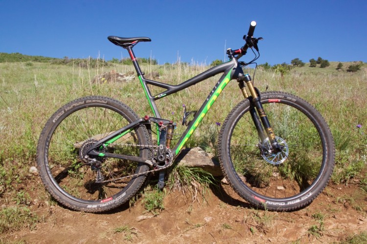 A mountain bike with a sleek, black and green frame is positioned on a patch of dirt surrounded by grasses and wildflowers. The bike features wide tires and a front suspension fork, set against a clear blue sky in a natural outdoor setting.