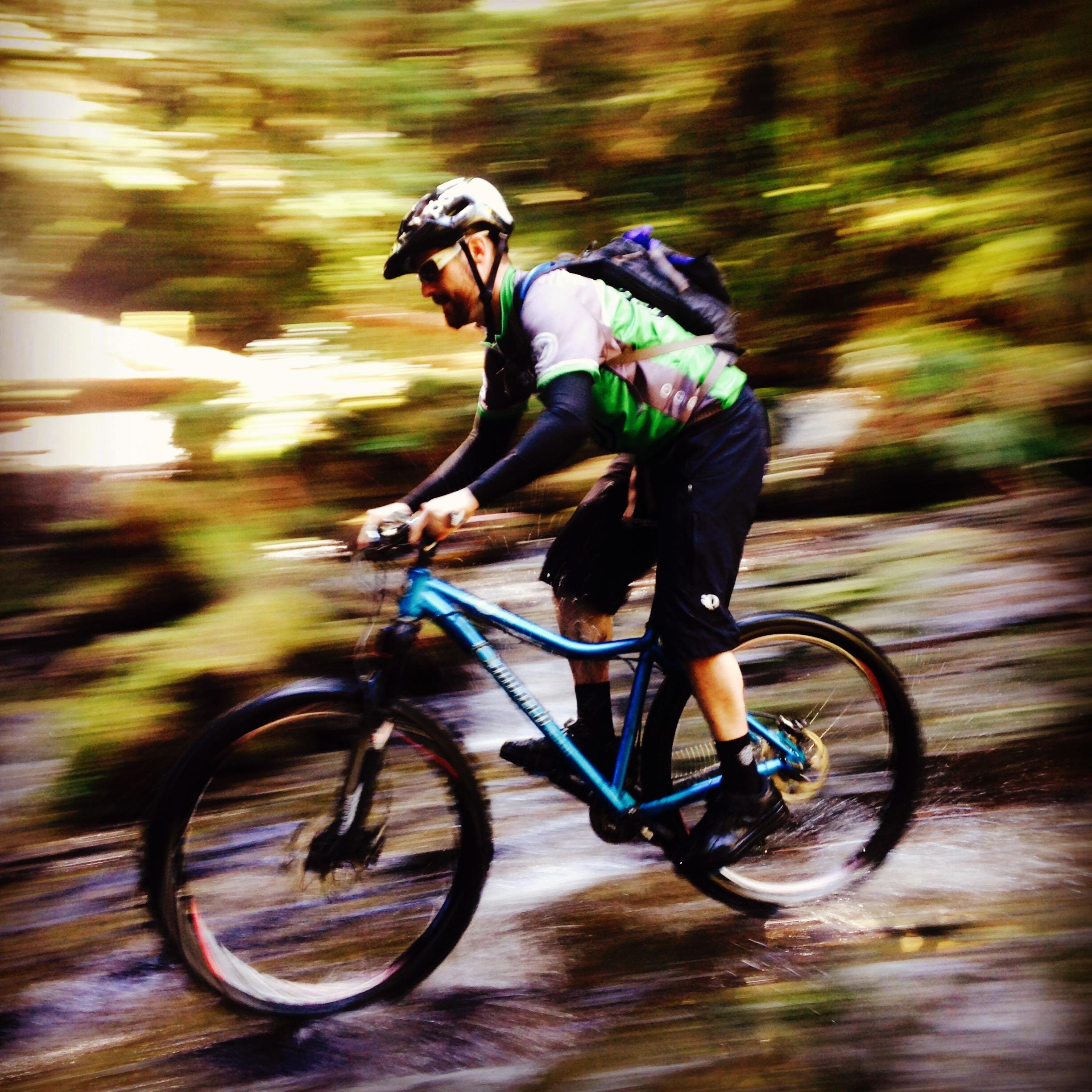 Canfield Brothers Yelli Screamy: A mountain biker in a green and gray jersey rides through a shallow stream, creating splashes as he pedals. The background is blurred, suggesting high speed and movement through a forested area. The cyclist wears a helmet, sunglasses, and a backpack, emphasizing an active outdoor adventure.