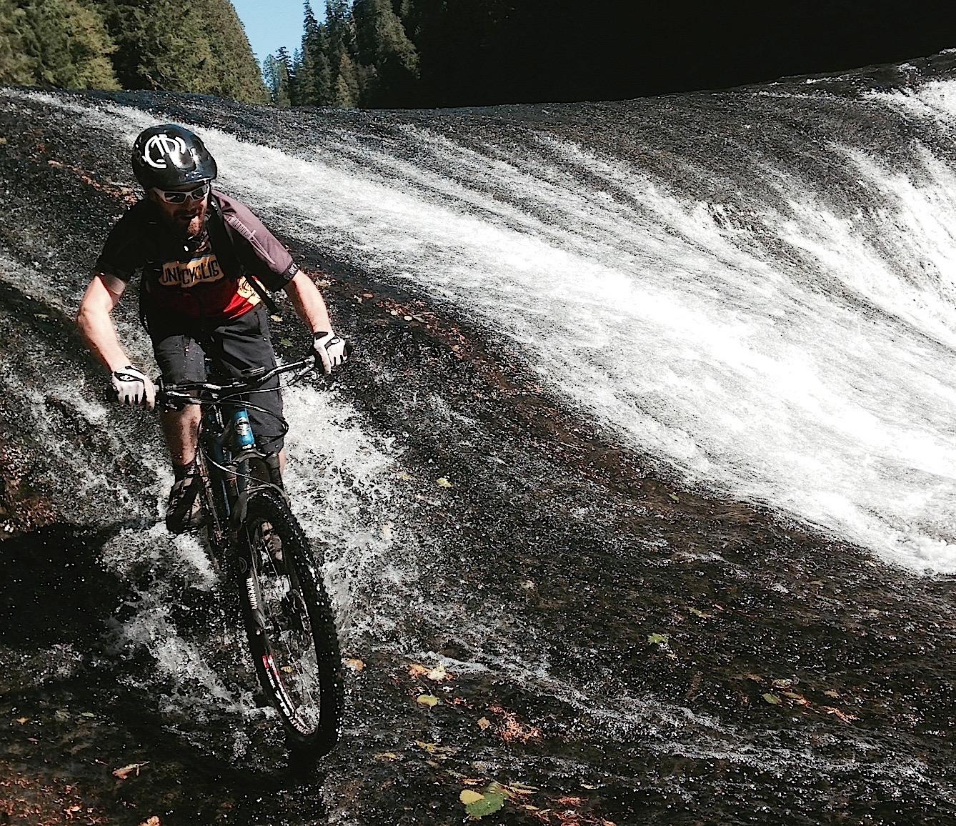 Canfield Brothers Yelli Screamy: A mountain biker navigating a rocky surface near a waterfall, splashing through water as he rides downhill amidst a forested landscape.