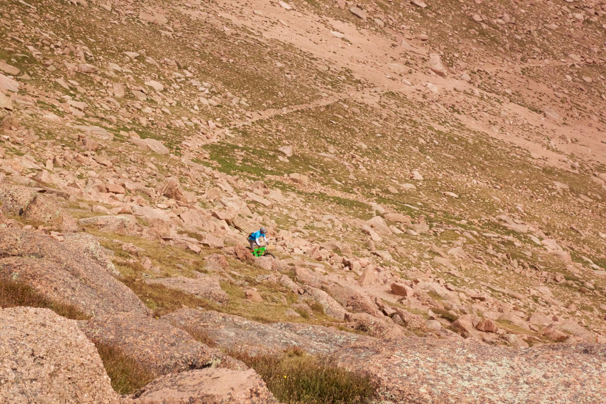 A person in a blue jacket is climbing a rocky slope in a mountainous landscape, surrounded by boulders and sparse vegetation. The terrain is steep and consists of brown rocks and patches of grass, with a trail visible in the distance. Barr Trail / Pikes Peak mountain bike trail.
