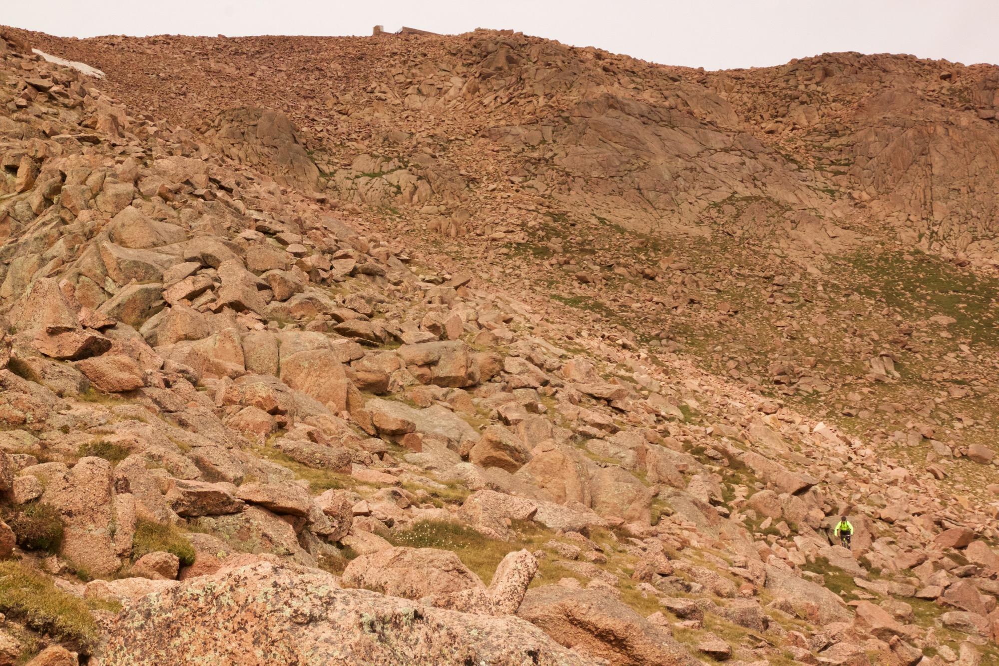 A rugged mountain landscape covered in rocky terrain, with a person wearing a bright yellow jacket walking amongst the boulders. The background features steep, rocky slopes and minimal vegetation. Barr Trail / Pikes Peak mountain bike trail.