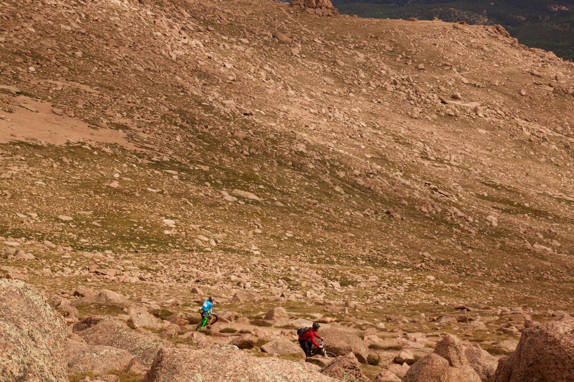 Two mountain bikers navigate a rocky, uneven terrain in a mountainous area. The landscape features large boulders and patches of grass, with a steep slope in the background. The sky is clear, suggesting a sunny day for outdoor activities. Barr Trail / Pikes Peak mountain bike trail.