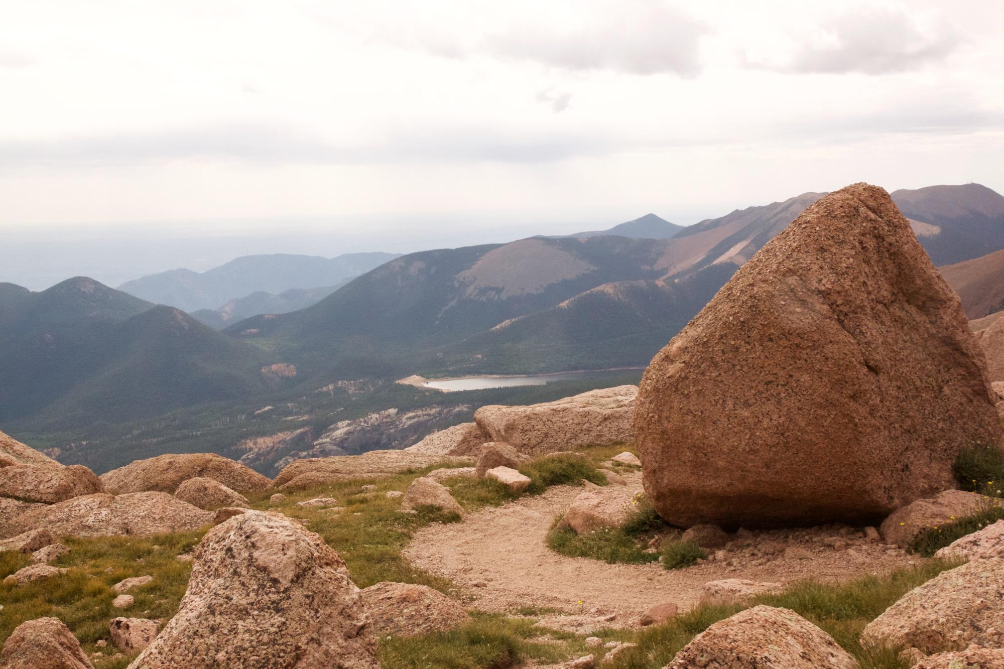 A panoramic view from a mountain summit, showcasing rocky terrain in the foreground and a range of green hills and mountains in the background under a cloudy sky. A small body of water can be seen nestled among the hills. Barr Trail / Pikes Peak mountain bike trail.