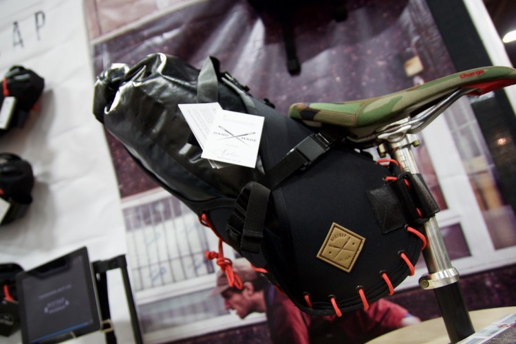 A close-up image of a black bike bag attached to a bicycle seat, featuring a shiny black waterproof material and orange accents. A camo-patterned seat cover is visible above the bag, along with a tag that reads "HANDMADE." The background is slightly blurred, showcasing other bags displayed at a trade show.