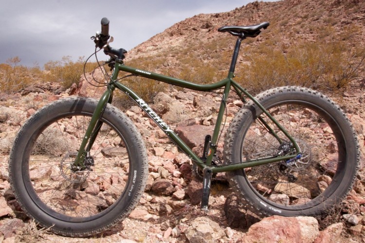 A green fat bike is parked on rocky terrain, showcasing its oversized tires and sturdy frame. The background features a rugged landscape with mountains under a cloudy sky, suggesting an adventure-ready setting.
