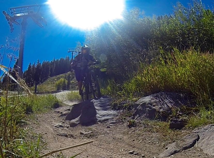 A mountain biker navigating a rocky trail in a sunlit landscape, with chairlifts visible in the background and lush greenery surrounding the path. Vail Mountain Bike Park mountain bike trail.