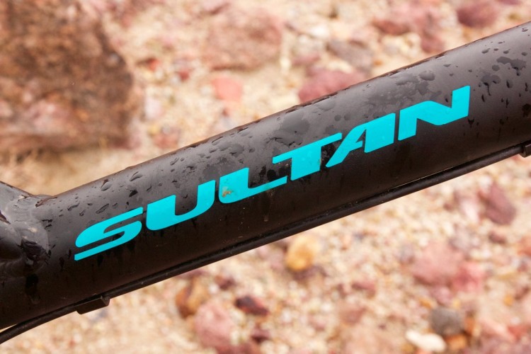Close-up of a black bike frame featuring the word "SULTAN" in bold aqua letters, with droplets of water on its surface, set against a background of rough terrain.