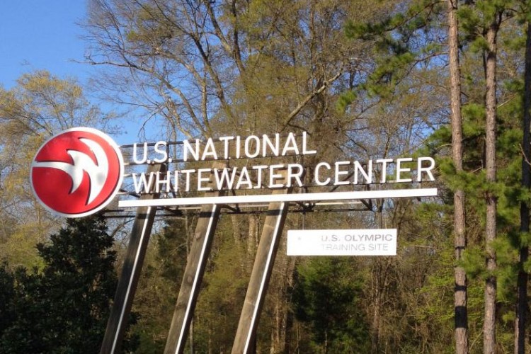 Sign for the U.S. National Whitewater Center, featuring the logo and text indicating it is a U.S. Olympic training site, set against a backdrop of trees.