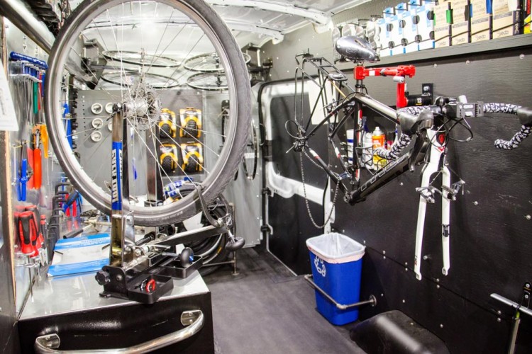 A well-equipped bike repair van interior featuring a bicycle wheel mounted on a stand, multiple bicycles hanging from hooks, and an array of tools organized on the walls. The space includes a workbench with various tools and manuals, as well as a trash bin in the corner.