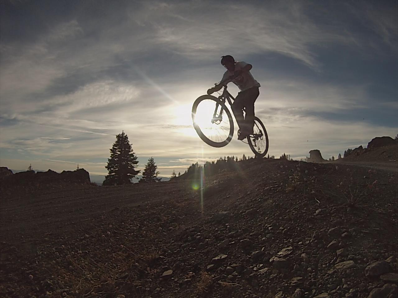 A mountain biker performing a jump on a dirt trail, silhouetted against a sunset sky with scattered clouds and distant trees. Anthony Peak Ridge mountain bike trail.