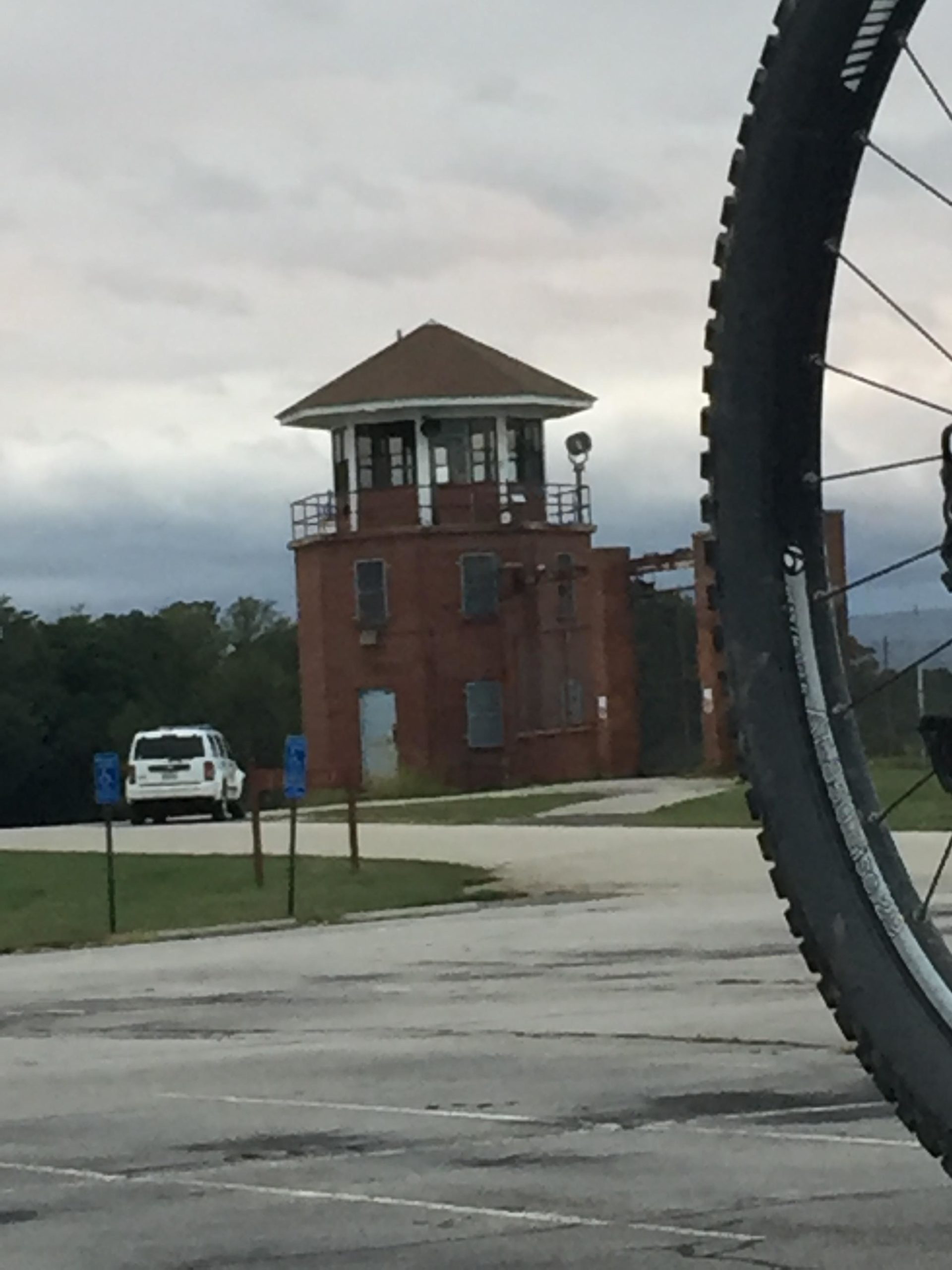 A historic red-brick building with a lookout tower in the background, partially obscured by the wheel of a bicycle in the foreground. A white vehicle is parked in a nearby lot, under a cloudy sky. Laurel Hill Park mountain bike trail.