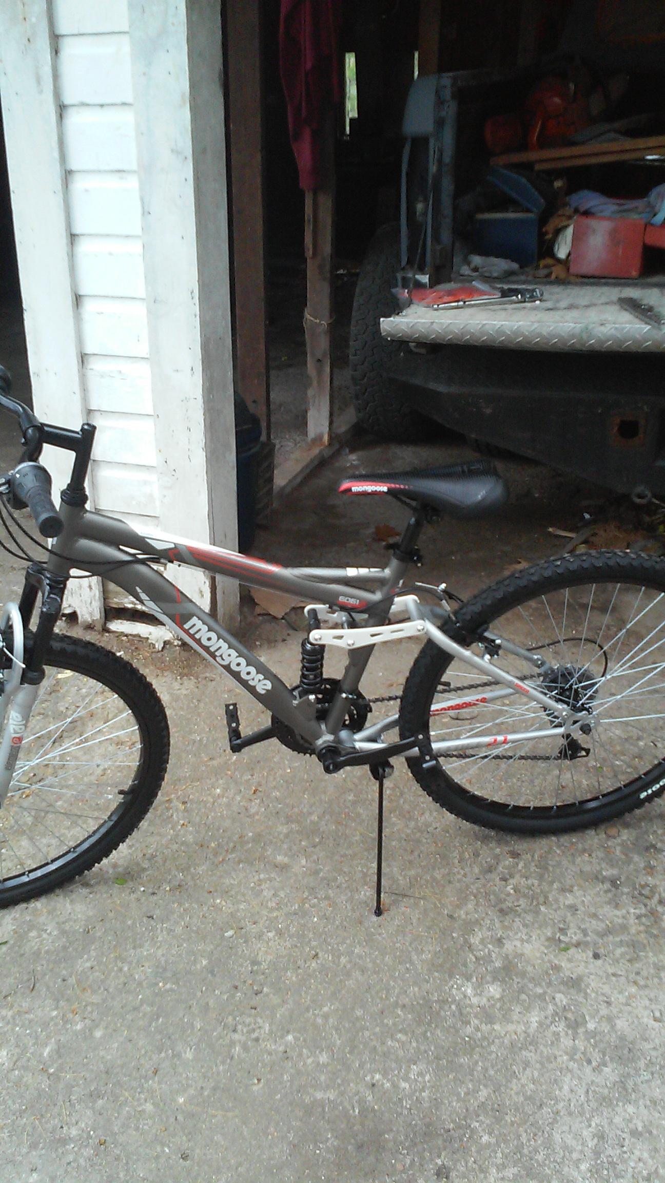 Mongoose Mongoose Ledge 2.1: A gray and red Mongoose mountain bike is parked on a gravel surface near a wooden structure. The bike features front suspension, a black seat, and prominent tires designed for off-road use. In the background, parts of a truck and various tools are visible, suggesting a garage or workshop setting.