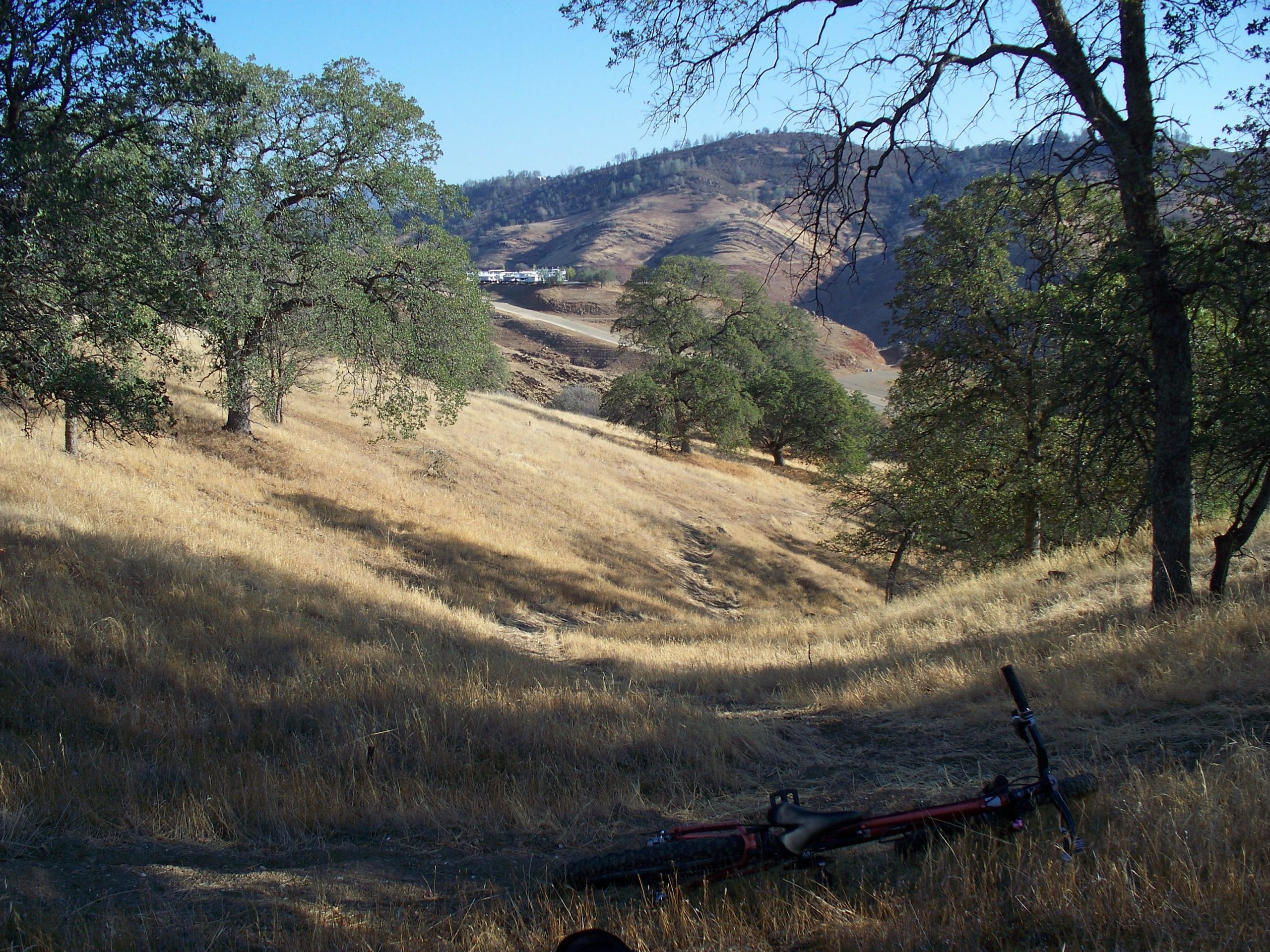 A serene landscape featuring a dry, grassy hillside with scattered trees. In the foreground, a red bicycle lies on the ground amidst the golden grass. In the background, rolling hills and a clear blue sky create a peaceful outdoor scene. Exchequer Mountain Bike Park mountain bike trail.