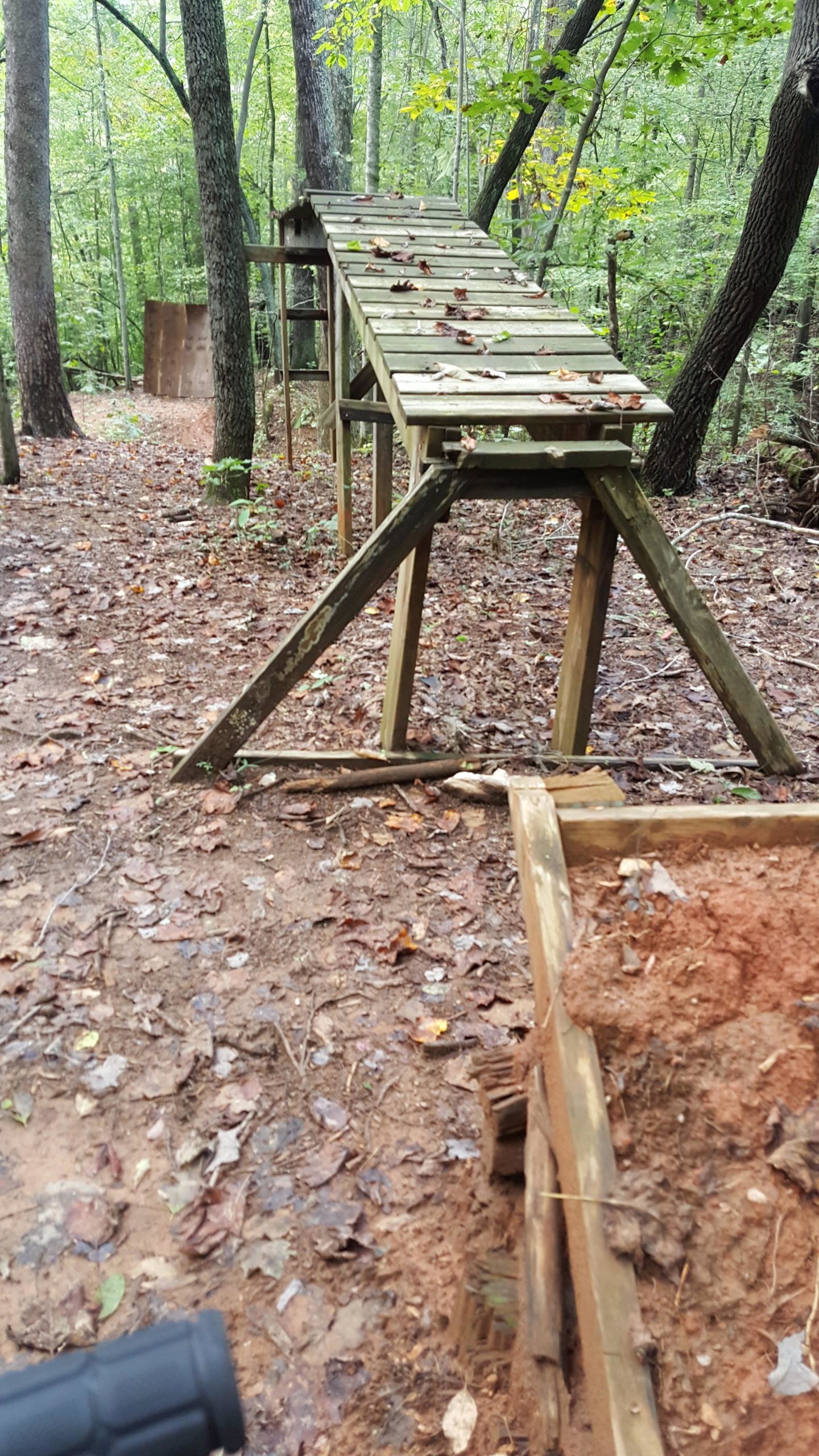A wooden ramp made from planks leads down into a forested area, surrounded by trees with yellow and green leaves. The ground is covered with damp fallen leaves and dirt, and there is a dirt ramp visible in the background. In the foreground, part of a wooden structure is partially visible, showing earth and debris. Salem Lake mountain bike trail.