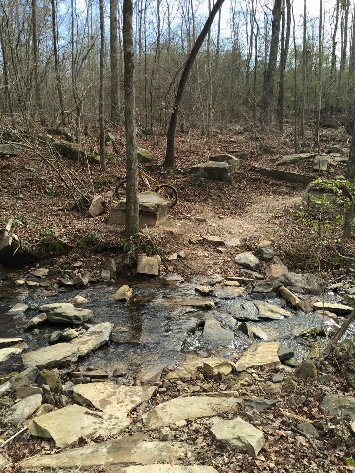 A rocky trail winding through a wooded area, bordered by trees and scattered leaves, with a small stream flowing across the path. A bicycle is leaning against a boulder in the background. Duck River mountain bike trail.