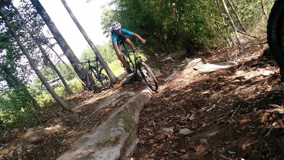 A mountain biker navigates a rocky trail surrounded by trees, showcasing an action shot of the rider leaning into a turn. Two bicycles are visible in the background, illustrating an outdoor biking adventure. The scene captures the rugged beauty of a forested area, with the ground covered in leaves and gravel. Duck River mountain bike trail.